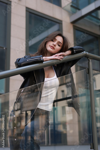 A beautiful brunette girl in a leather jacket, jeans and sneakers on the street of a modern city.