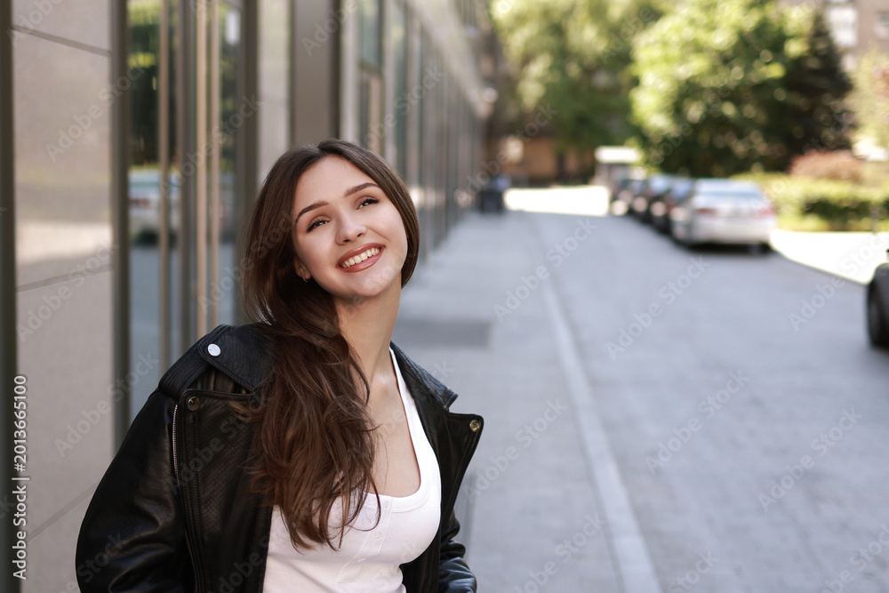 Fototapeta premium A beautiful brunette girl in a leather jacket, jeans and sneakers on the street of a modern city.