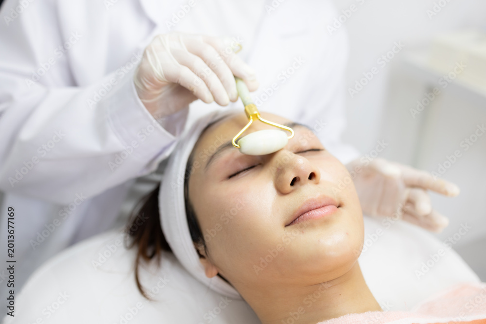 Naklejka premium Soft jade tool glides over calm woman's face. Gloved hands work gently with care and precision. Woman rests eyes closed, enjoying serene facial treatment. Gentle lighting enhances peaceful 