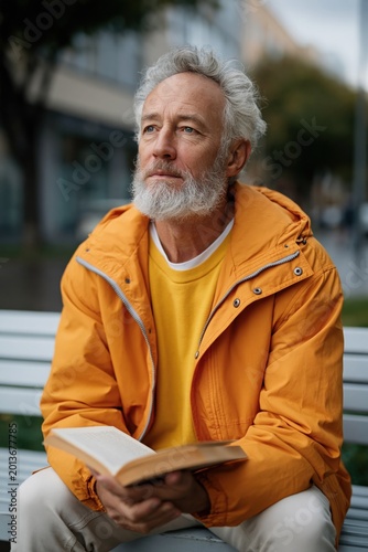 Senior man in orange jacket reading book on city bench