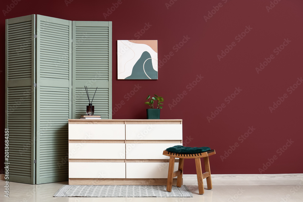 Fototapeta premium Interior of living room with folding screen, chair and home decor on commode near burgundy wall