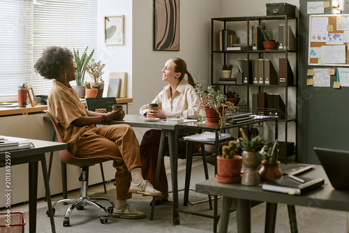 Fotografía African American young adult woman and Caucasian young adult woman meeting at office desk, discussing project over coffee