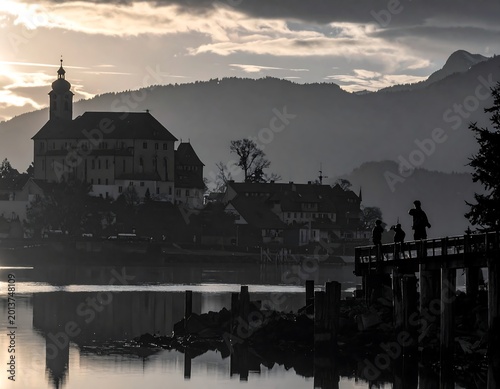 Silhouetted figures admire a lakeside town with a church under a moody sky, reflecting in still water