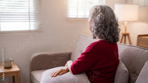 Photos Thoughtful elderly woman with gray hair sits alone on a cozy sofa, gazing out a bright window at home