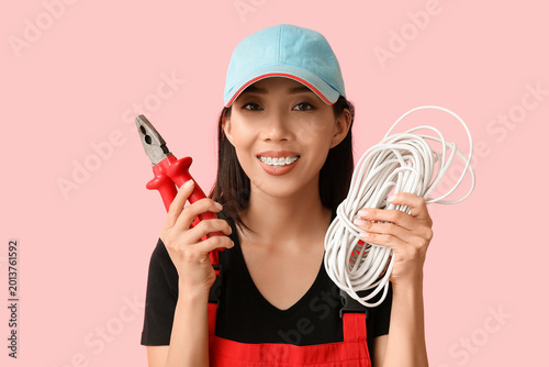 Young female electrician with pliers and cables on pink background