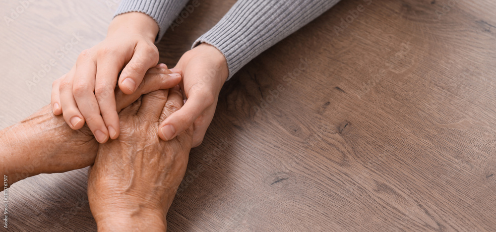 Fototapeta premium Young woman with grandmother holding hands on table, closeup. Care concept