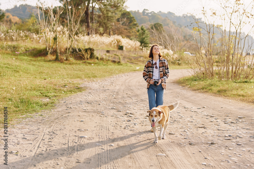 Obraz premium A woman and her dog walk along a dirt path in a sunny countryside setting, wearing a plaid shirt and jeans, as they move through an open field and trees in a peaceful outdoor moment.