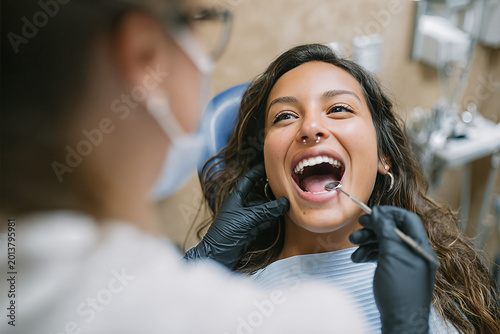 Young woman patient with septum ring laughing wide as dental hygienist in black gloves uses a mouth mirror during a bright community clinic exam