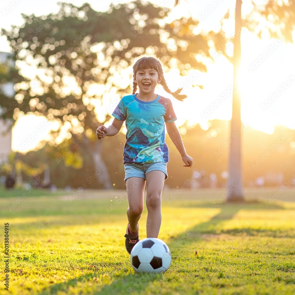 Obraz premium Joyful young girl with braids plays soccer in a sunlit park during golden hour. This energetic shot captures childhood happiness and healthy lifestyle, perfect for family or sports ads.
