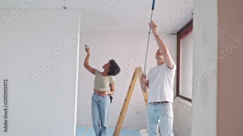 A couple diligently paints a room's ceiling during their home renovation project. The woman focuses on a spot on the wall, while the man, on a small step ladder, stretches to paint higher areas.