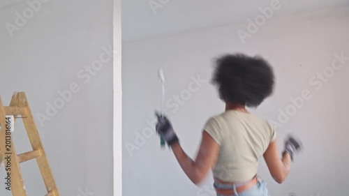 A young woman with an afro hairstyle energetically paints a room's white wall, wearing gloves and a casual outfit, with a ladder and paint supplies nearby in a bright, minimalist space.