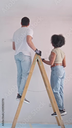 A couple paints a wall in a bright, minimalist space, the man on a wooden ladder carefully applies paint while the woman supports him from the ground.