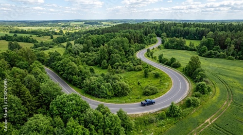 A dark blue car navigates a winding road through a lush green landscape. The road curves through a valley surrounded by dense forests and expansive fields.