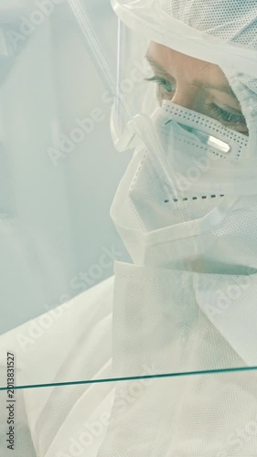 A young scientist in a white protective suit, mask, and visor examines a specimen in a sterile cleanroom, highlighting the precision and care of the medical laboratory.