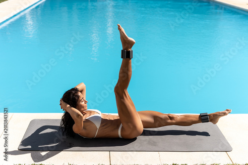 Fotografie Fit woman in white bikini performing leg raise crunch with ankle weights on mat beside pool, engaging abdominal muscles and demonstrating core strength during a bright outdoor summer fitness session