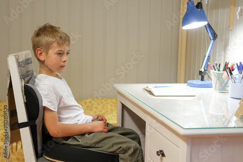 Foto Boy kid sitting at study desk under lamp, hesitant expression while facing blank notebook and pencil cup, tidy bedroom, evening light creates quiet atmosphere, preparing homework