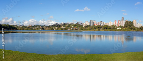 Barigui Park lake and island in Curitiba, Parana, Brazil, showcasing the modern city skyline against a bright blue sky.