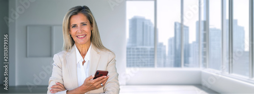 female real estate agent holds a phone and smiles in a bright, modern apartment with the city skyline in the background. Real estate professional