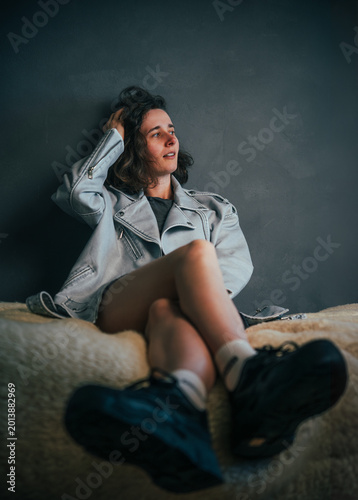 Beautiful young girl with beautiful legs adjusting her hair while seated against a textured wall in a relaxed pose, exuding a thoughtful expression in soft lighting and muted colors.