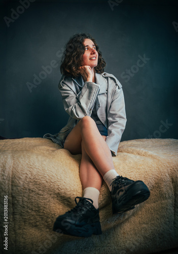 Beautiful young girl with beautiful legs exuding playfulness and joy, sitting comfortably on a bed in casual sporty attire, lost in thought within a warm and inviting atmosphere.