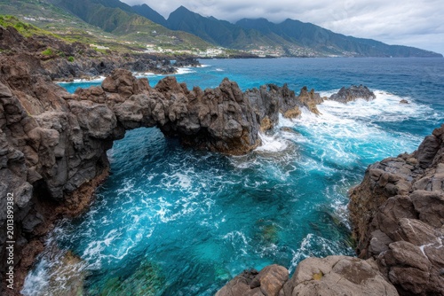 Waves crash against a rocky coastline under a cloudy sky with mountains visible in the background