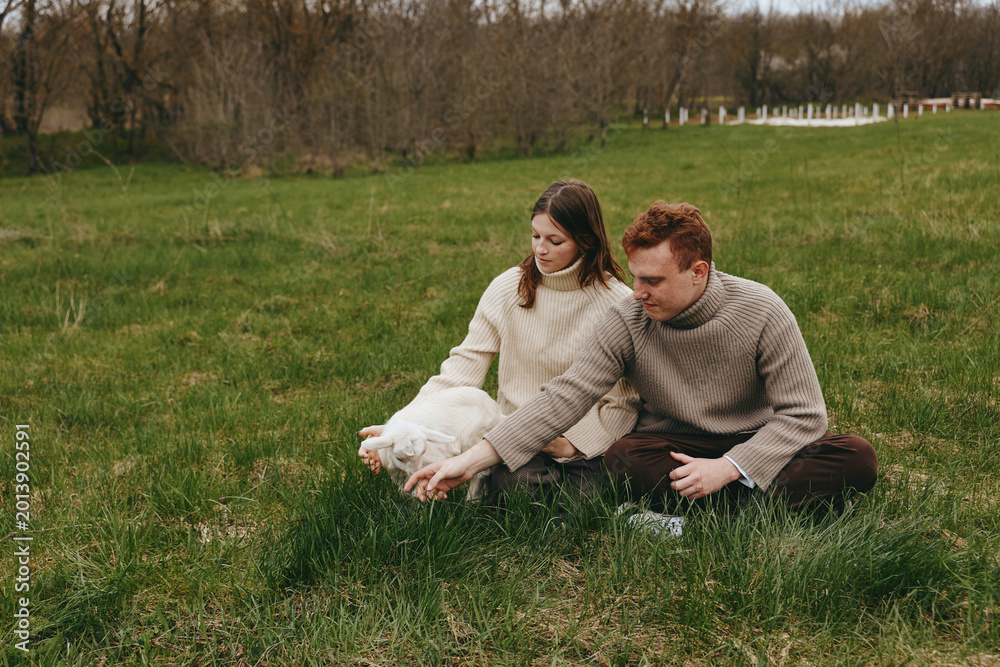 Fototapeta premium Lifestyle scene of a couple outdoors on a grassy field, smiling as they cradle a playful goat kid, cozy sweaters, natural light, gentle breeze, calm countryside atmosphere