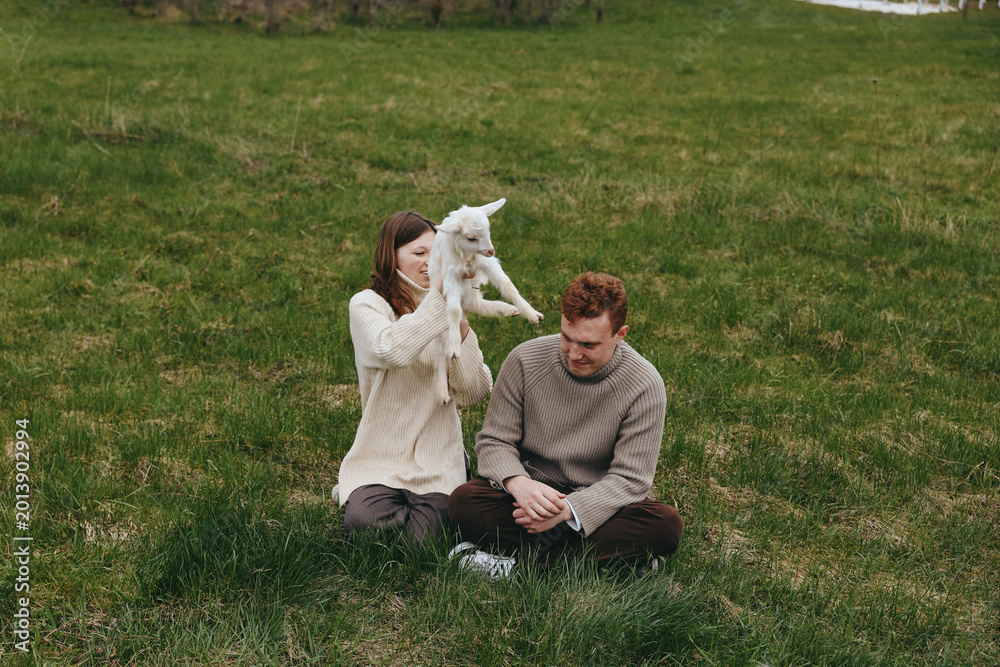 Fototapeta premium A couple sits on grass with a playful kid goat nearby, sharing a tender outdoor moment in a natural meadow, cozy sweaters, relaxed mood, and rustic countryside ambiance.