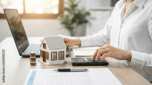 A person calculating property investments with a calculator, surrounded by coins and a house model, representing real estate business.
