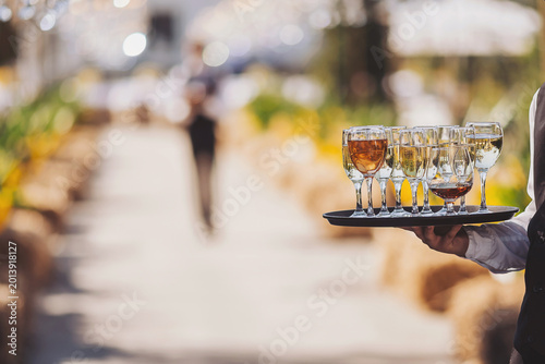 A waiter holds a tray filled with drinks in clear glasses while walking through a garden venue. Guests can be seen enjoying their time in the background