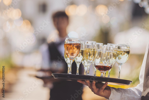 A waiter holds a tray with several glasses of different drinks. The setting is outdoors, with people socializing in the background. The mood is lively as guests enjoy the evening