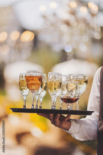 A waiter holds a tray filled with various glasses of wine at a gathering outside. The event takes place in a garden, and the sun shines bright in the background