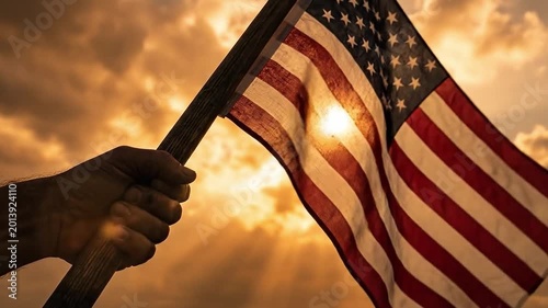 Close-up of strong hand holding American flag against glowing sunset sky, sunlight shining through fabric with dramatic rays creating emotional patriotic freedom concept in cinematic style.
