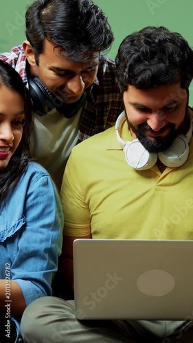 Indian college students using laptop while seating against green wall on floor indoors, discussing notes, researching online, learning together in friendly atmosphere, working on group project