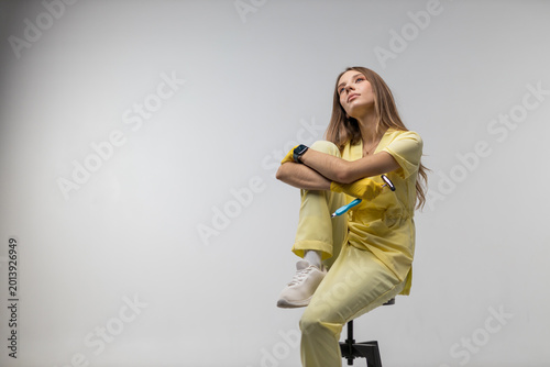Quadro su tela Female nurse in yellow medical scrubs seated on a stool looking thoughtful and u