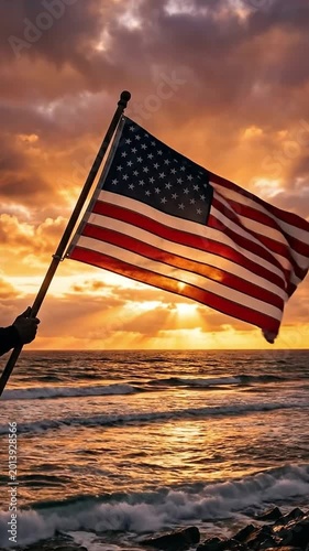 Male hand holding a large American flag above ocean waves during dramatic sunset with glowing clouds and golden sun rays, creating a powerful patriotic and cinematic atmosphere.

