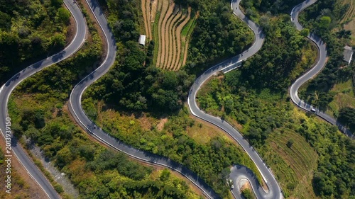 Top-down drone view of winding road in Ha Giang, Vietnam. Serpentine mountain route crossing green hills and terraced landscape with scenic curves from above.