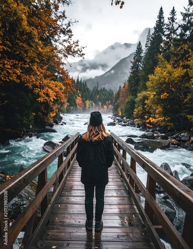 Woman In Black Jacket Standing On Wooden Bridge Overlooking Turquoise River And Misty Mountains In Autumn Forest Landscape Back View