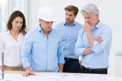Obraz Group of professionals analyzing architectural plans on a table in a bright office space, featuring a man in a hard hat, two men in blue shirts, and a woman in a white blouse
