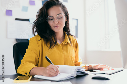 Concentrated attractive woman siting at office desk and writing plans in her notebook. Smart white woman office worker signs papers. Office job or freelance worker concept.