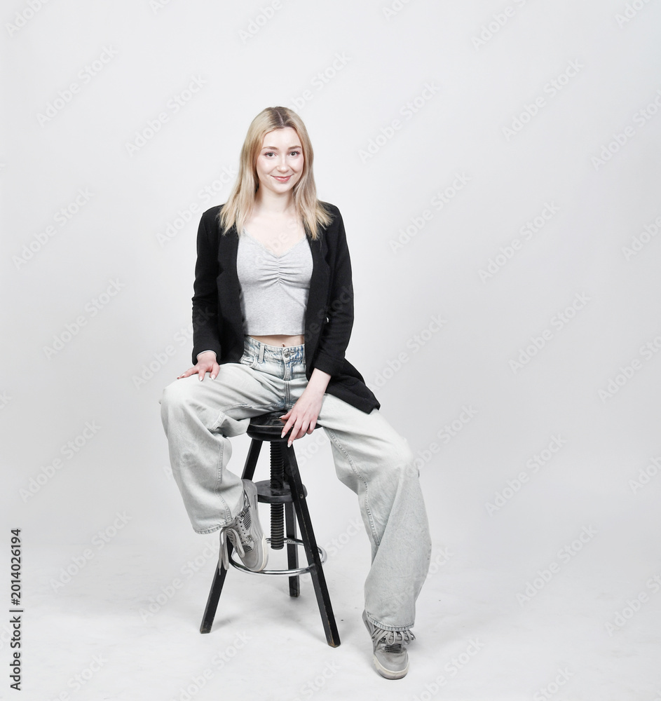 Fototapeta premium Junge blonde Frau sitzt entspannt auf Hocker vor hellem Studiohintergrund – modernes Lifestyle- und Fashion-Porträt mit natürlicher Ausstrahlung und viel copyspace und copy space.