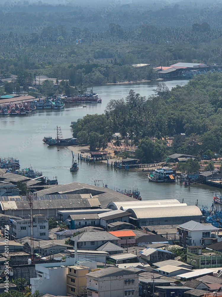 Fototapeta premium Aerial view showing calm peaceful coastal town with flowing river passing through industrial warehouse building and residential house near dense green forest under hazy daytime sky