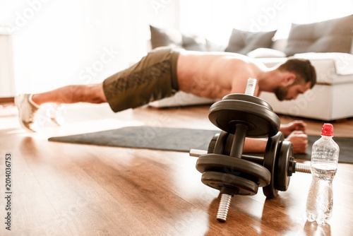 Big dumbbells on floor. Young man doing sport workout in room during quarantine. Side view t-shirtless guy sportsman stand on plank position. Straight well-built body.