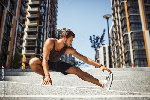 Young man exercising outside. Picture of strong guy stretching out his leg and holding it with hand. Standing alone in yoga position. Warming up before exercise or finishing training with stretching.