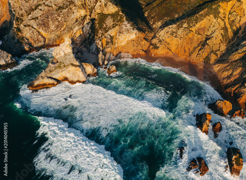 Fotografie Aerial view of dramatic rocky coastline with crashing ocean waves and golden sun
