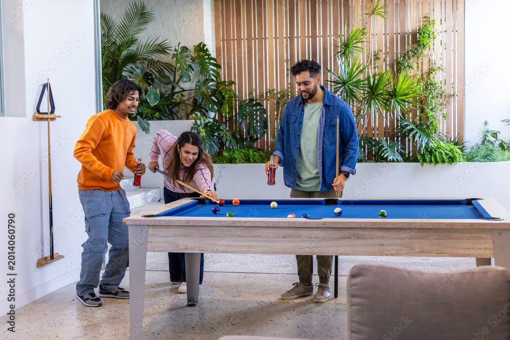 Fototapeta premium Diverse friends leaning, taking shots and chatting in lounge with blue-felt pool table and cues