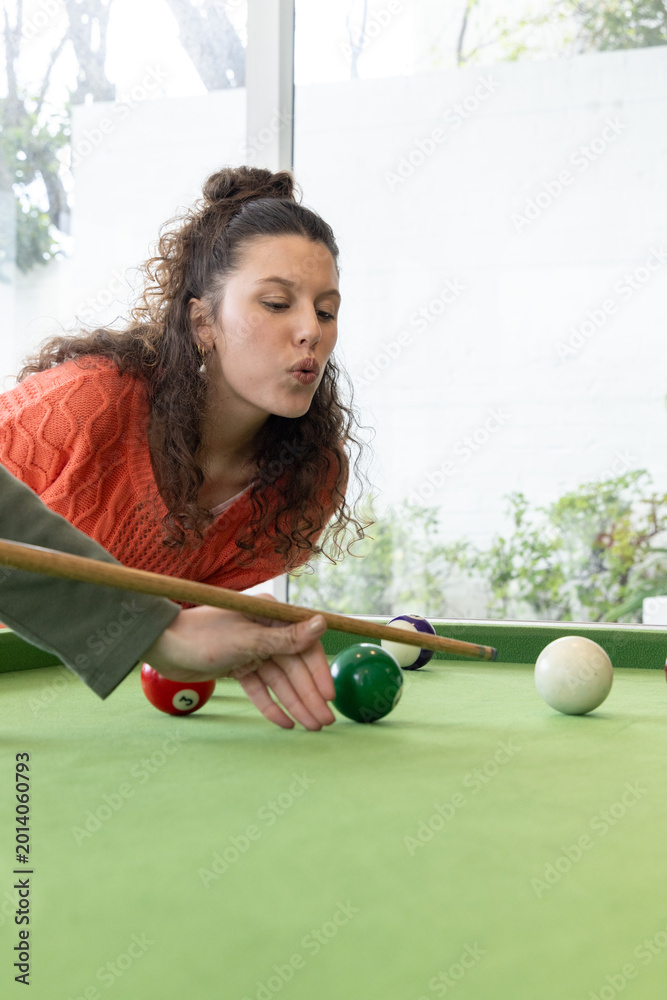 Fototapeta premium Adult female with partner's bridge leaning over pool table at home aiming cue in orange sweater