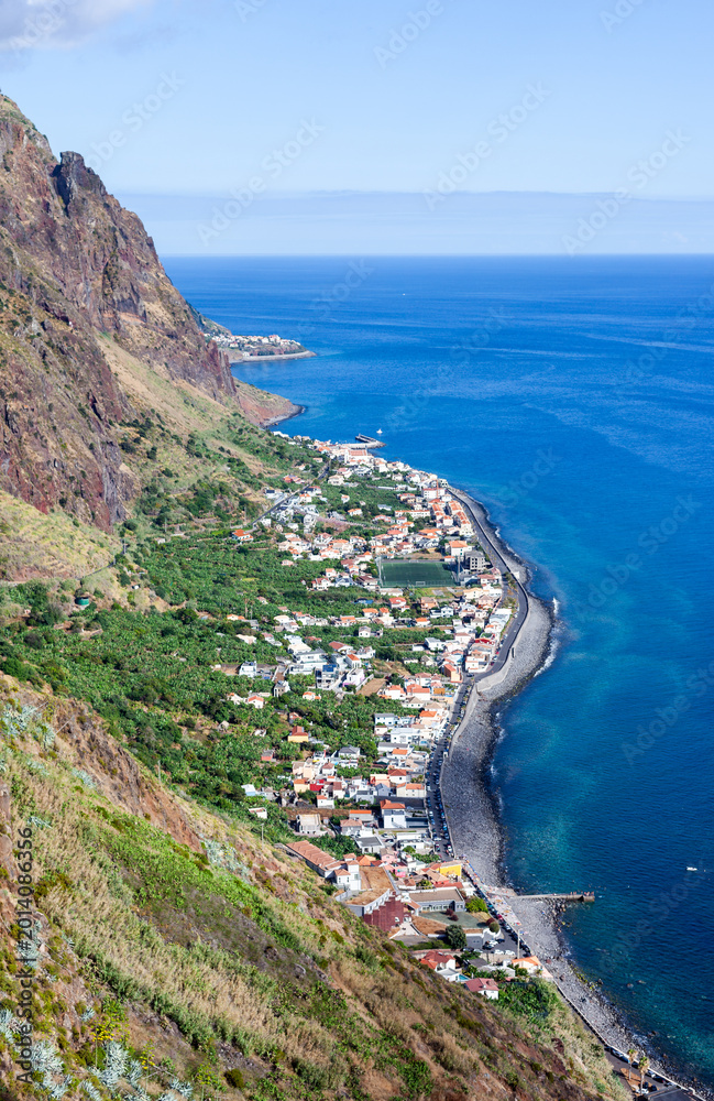Fototapeta premium Madeira coastal village. White houses cluster along a curved black pebble shoreline on northern Madeira. Steep cliffs rise behind the settlement with terraced green slopes.