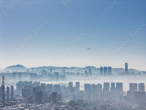 Aerial View of Modern Cityscape with High-Rise Buildings Emerging from Morning Fog and a Distant Airplane in the Sky
