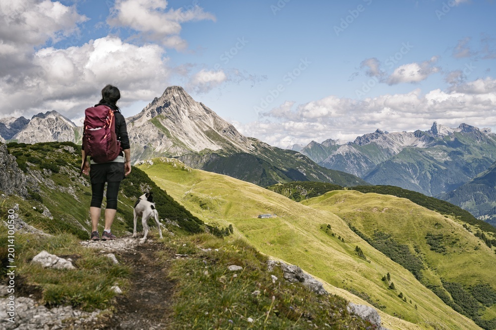 Fototapeta premium Female Hiker with Dog on Alpine Ridge Overlooking Biberkopf Mountain Peak in Bregenzerwald, Austria