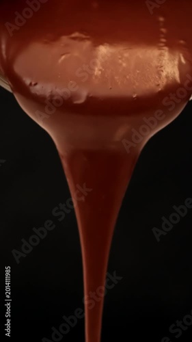 Glossy melted chocolate drips slowly from above in thick stream against dark background with smooth shine
